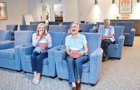 Three people are sitting in a room, enjoying a movie. They are in a row of blue chairs, with one person on the left, one in the middle, and one on the right. The person on the left is holding a red cup, while the person in the middle is holding a red popcorn bucket. The person on the right is also holding a red cup. AI generated content
