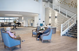 Two elderly women sit in chairs in a lobby, one on the left and one on the right. They are both wearing glasses. The room has a piano and a vase with flowers on a table. There are also two couches and a potted plant in the room. AI generated content